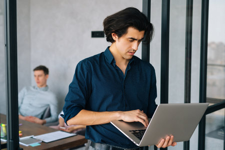 Portrait of concentrated male entrepreneur in casual clothes using typing laptop holding in hand standing in office, looking to screen. Startup team discussing project sitting at desk on background.の写真素材