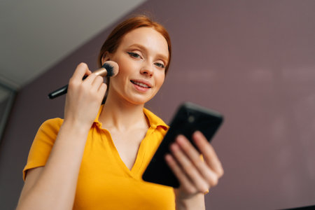 Closeup low-angle view of pretty young woman applying cosmetic powder with big professional makeup brush and using smartphone at home, smiling looking at camera. Attractive female using blusher.の写真素材