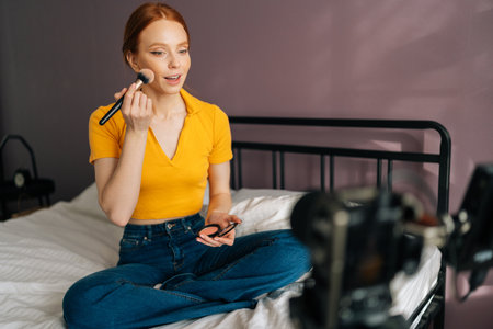 Pretty beauty female blogger recording cosmetics product review, applying blush on cheek, recording video for channel using professional camera standing on tripod, sitting on bed in home studio.の写真素材