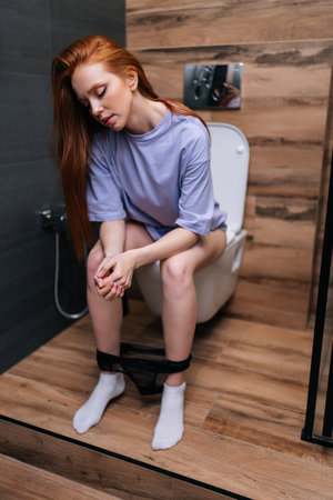 Vertical portrait of bored young woman in casual clothes sitting on toilet bowl at home bathroom with modern interior. Tired exhausted redhead female sitting in restroom after party feeling sleepy.の写真素材