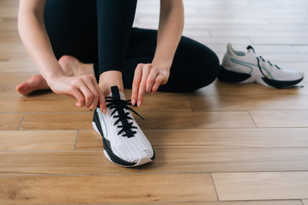 Closeup cropped shot of unrecognizable athletic female tying shoelaces on sports shoe preparing to workout indoors. Close up hands of sporty woman tying shoe laces on floor preparing for workoutの写真素材