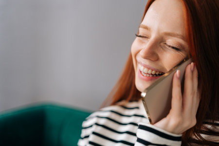 Close-up face of laughing young woman talking on smartphone at home. Cheerful redhead female making answering call by cellphone. Happy pretty lady having pleasant funny conversation speaking by mobileの写真素材