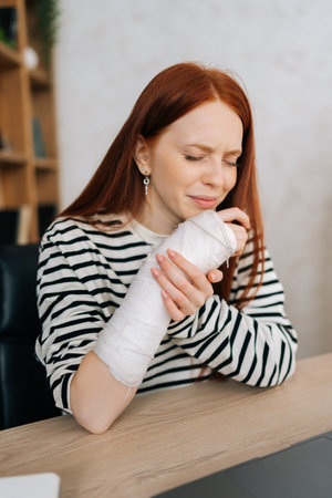 Vertical portrait of suffering from pain crying young woman with broken right hand wrapped in gypsum bandage sitting at table closed eyes. Sad redhead female with broken arm from accident at home.の写真素材