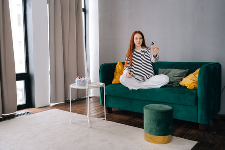 Wide shot of suffering from pain sad female vlogger with broken arm wrapped in white gypsum bandage holding smartphone talking to camera shooting vlog, making video call at home, sitting on sofa.の写真素材