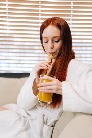 Vertical portrait of pretty young woman in white bathrobe drinking cocktail from straw with closed eyes sitting on sofa by window at spa resort. Charming female relaxing after body treatment.の写真素材