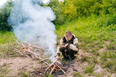 Front view of frozen survivalist male in raincoat putting firewood on campfire to making fire on bank of river in evening before sunset. Concept of exploration, travel and adventure.の写真素材