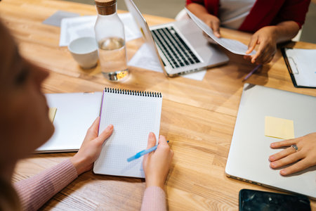 Close-up high-angle view of focused businesswoman holding notebook and pen in hand and write during multicultural professional businesspeople working together, on research plan in boardroom.の写真素材