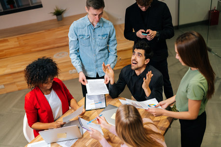 High-angle view of frustrated team leader screaming in desperation at multiethnic corporate meeting at desk. Exhausted businessman feeling tired at briefing with diverse male and female colleaguesの写真素材