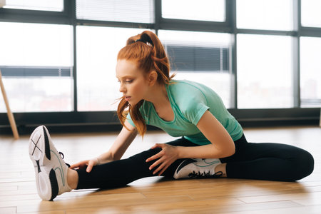 Focused flexible fit woman in sportwear practicing head to knee forward bend asana in yoga studio. Sporty redhead female relaxing in deep stretching in Janu Sirsasana. Concept of healthy lifestyle.の写真素材
