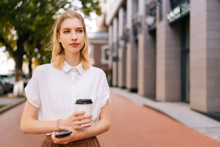 Portrait of confident blonde lady in casual outfit holding in hands mobile phone and takeaway coffee pensive looking away while standing outside coffee shop, on city street on summer sunny day.の写真素材