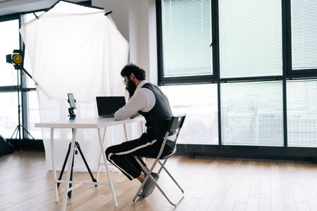 Remote rear view of Indian social media influencer recording video blog podcast on smartphone sitting at table with professional lighting equipment. Blogger talking to camera record video by window.の写真素材