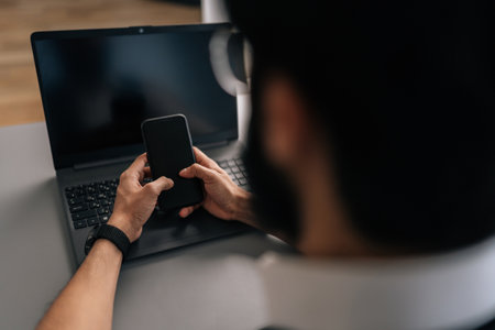 Closeup view from back of unrecognizable Indian businessman sitting at desk using modern laptop and smartphone gadget. Male manager consult client work on computer and cellphone device at workplace.の写真素材