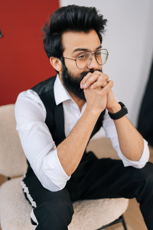 Vertical portrait of bearded gentleman wearing elegant formalwear sitting on chair and confidently looking away. Front view of handsome Indian young man in fashion glasses posing at studio.の写真素材