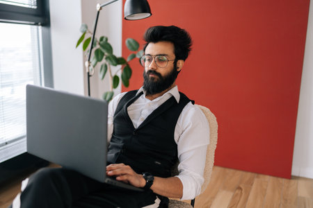 Portrait of Indian business man in formalwear working on laptop sitting soft poof in light coworking office on background of window. Bearded freelancer male in eyeglasses looking at computer screen.の写真素材