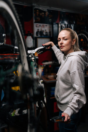 Vertical portrait of attractive bike mechanic female repairing and fixing mountain bicycle standing on bike rack in repair workshop with dark interior, looking at camera. Concept of bike maintenance.の写真素材