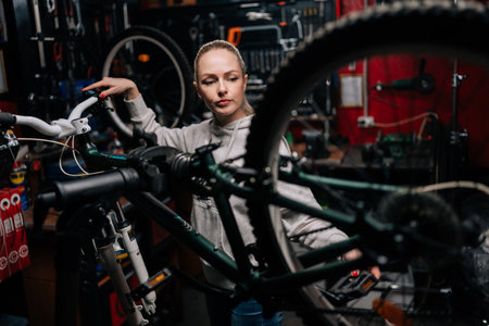Focused cycling mechanic female watching rear shifter of mountain bike, changing speeds using handlebar shift lever, working in bike repair workshop with dark interior. Concept of bike maintenance.の写真素材