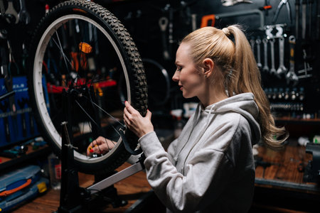 Side view of smiling blonde female cycling craftsman checking bicycle wheel spoke with bike spoke key in repair workshop with dark interior. Concept of professional maintenance of bicycle transport.の写真素材