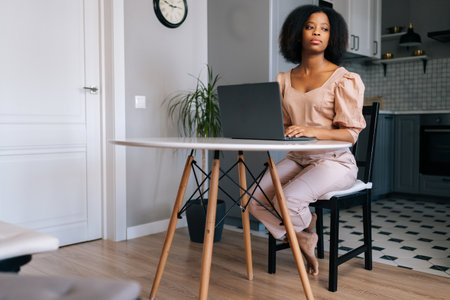 Thoughtful African American female freelancer programmer working remotely from home office using laptop siting at desk in kitchen pensive looking away. Motivated black female enjoying freelance jobの写真素材