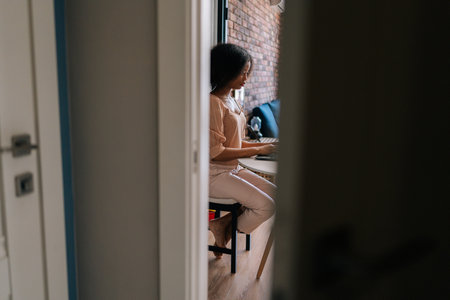 Remote side view of curly African female student typing on notebook keyboard studying working online. Black businesswoman working on laptop at desk. Afro lady working remotely from home office.の写真素材