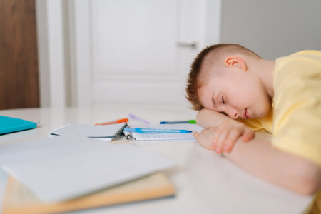 Close up side view of exhausted pupil boy sitting at home or classroom lying on desk filled with books training material and notebook. Tired schoolchild sleeping lazy bored to study lack of energy.の写真素材