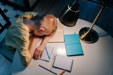 Top view of tired little schoolboy sleeping at table with books, copybook and textbook under light...