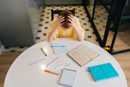 Top view of exhausted pupil boy tired from studying holding head head with hands sitting at desk...