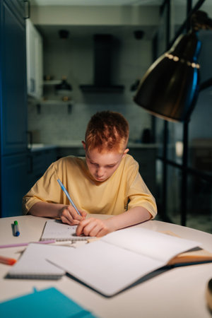 Vertical portrait of pupil student boy studying at home writing in exercise book doing homework, learning sitting at table under light of lamp at night. Schoolboy reading textbook sitting at desk.の写真素材