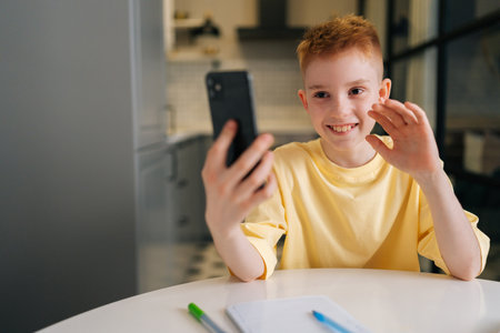 Portrait of happy child schoolboy waving hand, looking at smartphone distance learning class using...