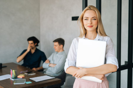 Portrait of friendly female entrepreneur in casual clothes holding laptop in hand standing in office, looking at camera. Startup business team discussing project sitting at desk on background.の写真素材