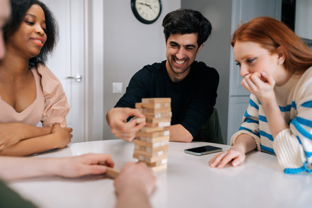 Cheerful young man carefully pulling out wood cube from wooden tower structure sitting at table....