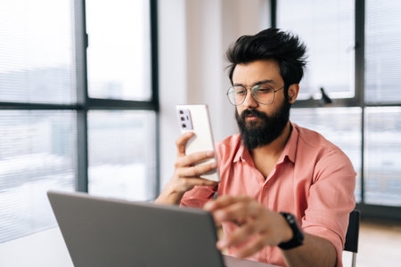Portrait of bearded Indian businessman in glasses holding mobile phone texting online message...