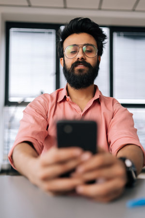 Vertical portrait of Indian bearded businessman in eyeglasses using smartphone sitting at office desk by window. Selective focus shot of handsome freelancer texting message on mobile phone.の写真素材