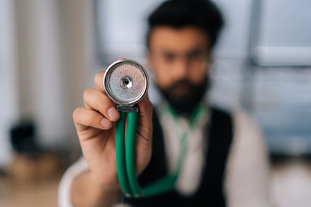 Closeup selective focus of blurred Indian male cardiologist in glasses holding stethoscope on hand for listening check up lungs or heart. Medium shot of doctor using stethoscope looking at camera.の写真素材