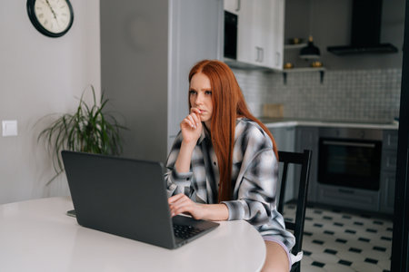 Portrait of thoughtful young woman sitting at table with laptop computer, pensive looking away, touching chin in kitchen. Serious redhead female freelancer remote working on laptop from home office.の写真素材