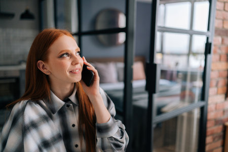 Close-up face of cheerful young woman holding mobile phone happily calling and chatting at home, smiling looking away. Closeup of laughing redhead female having pleasant conversation on smartphone.の写真素材