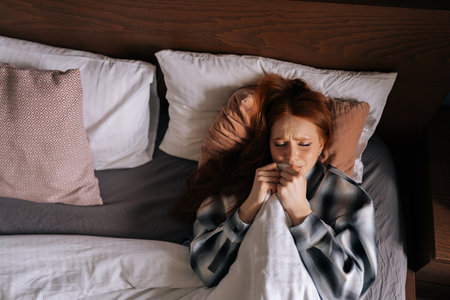 Top view portrait of sad depressed young woman crying hiding under blanket lying on bed in dark bedroom. Lonely upset beautiful female lying alone in bed, suffering from heavy thoughts.の写真素材