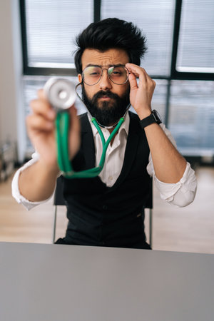 Vertical portrait of professional Indian male doctor in glasses holding stethoscope on hand for listening check up lungs or heart. Front view of practitioner using stethoscope looking at camera.の写真素材