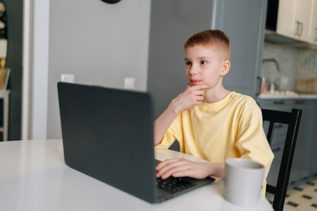 Portrait of pensive schoolboy typing on laptop computer and thinking with hands on chin, pensive looking away. Pupil child boy learning lesson at school, making homework and using notebook gadget.の写真素材