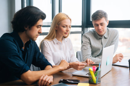Close-up of successful young male and female startup business colleagues having discussion sitting at desk looking at laptop screen in light office room by window. Concept of corporate work meeting .の写真素材