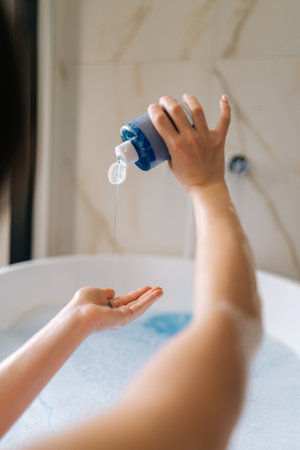 Vertical back view of unrecognizable young woman taking bubble bath, pouring shampoo into hand,...
