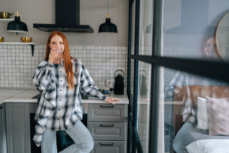 Portrait of happy thirsty young woman holding glass, make sip, drinking still water, enjoying natural aqua standing in home kitchen, preventing organism dehydration, smiling looking away.の写真素材