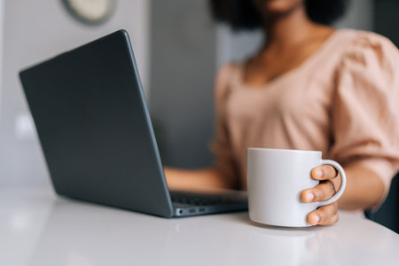 Close-up cropped shot of unrecognizable black female freelancer typing on laptop computer and holding cup with coffee or tea in morning sitting at table, working from remote home office workplace.の写真素材