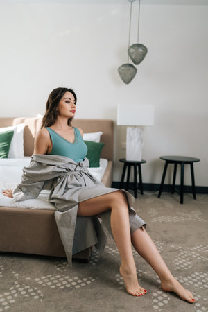 Vertical side view of seductive brunette female in revealing robe and bra sitting on bed in luxury bedroom in sensual erotic posture, eyes closed. Sexy young woman in gorgeous underwear posing at homeの写真素材