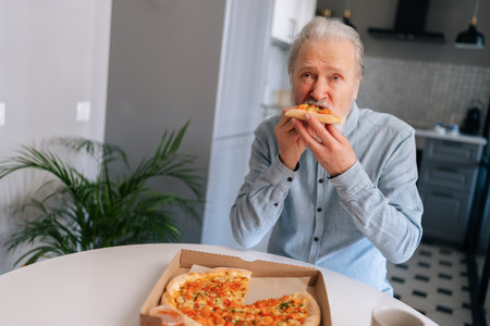 Vertical portrait of hungry gray-haired mature male enjoying to eat pizza alone sitting at kitchen table. Front view of bearded senior older man with appetite to eat tasty pizza at home.の写真素材