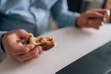 Detail cropped shot of unrecognizable senior older businessman with wrinkle hands eating hamburger with beef from fast food restaurant sitting at table, working online with laptop computer.の写真素材