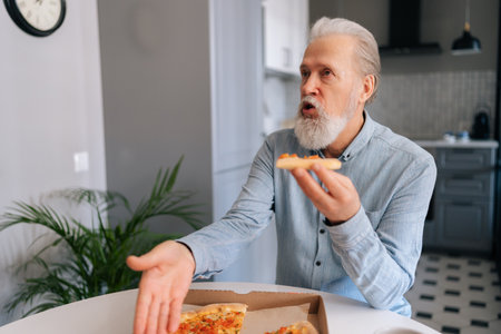 Angry annoyed bearded mature elderly male tasting slice of pizza with suspicion and with grimace of displeasure putting aside bad quality food sitting at table in home kitchen, close-up, cropped shot.の写真素材
