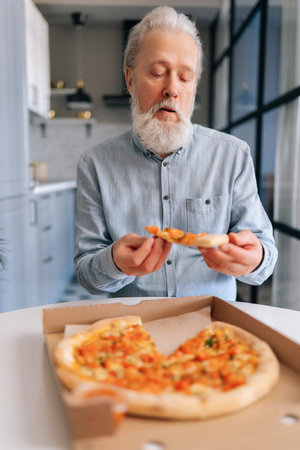 Vertical portrait of hungry senior older man with grey hair enjoying to eat pizza alone sitting at kitchen table. Front view of bearded mature elderly male with appetite to eat tasty pizza at home.の写真素材