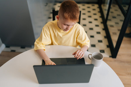 Top view of cute redhead 10 years old boy looking to laptop screen and typing doing homework sitting...