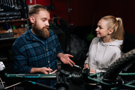 Front view of bearded cycling repairman male having conversation with blonde female client, talking about problem of bicycle detected during diagnostics, in repair shop with dark interior.の写真素材