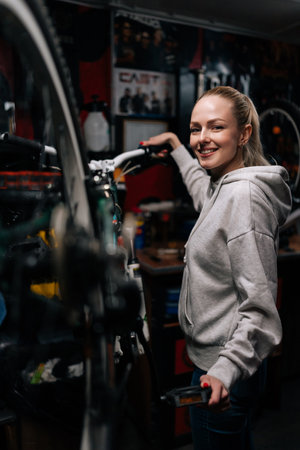 Vertical portrait of cheerful cycling mechanic female repairing and fixing mountain bicycle standing on bike rack in bike repair workshop with dark interior, smiling friendly looking at camera.の写真素材